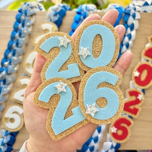 May include: A hand holds a set of light blue and gold glitter graduation year cutouts with star accents. The numbers "2026" are displayed. Other graduation leis are visible in the background, with blue and white ribbons.