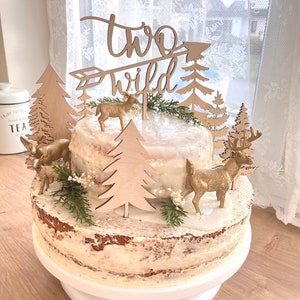 Könnte beinhalten: Eine zweistöckige Torte, verziert mit einem Holz-Topper 'two wild', goldenen Hirschfiguren und Holzbäumen. Die Torte ist mit einem weißen Buttercreme-Frosting überzogen und hat einen rustikalen, natürlichen Look.