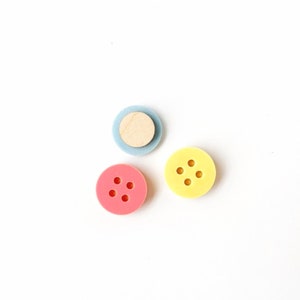 Op de afbeelding: Drie knoppen, één lichtblauw, één roze en één geel, elk met vier gaten. De lichtblauwe knop heeft een houten cirkel in het midden.