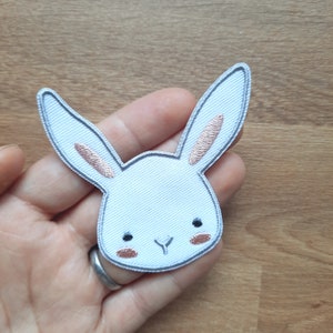 Könnte beinhalten: Ein weißer gestickter Hasen-Patch mit rosa Ohren. Der Hase hat ein einfaches, cartoonartiges Gesicht mit schwarzen Augen und einer rosa Nase.