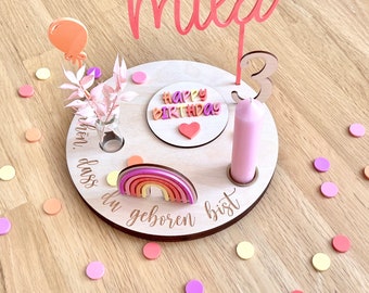 May include: A wooden birthday cake stand with a pink candle, a rainbow, and the words "Happy Birthday" and "Schon dass du geboren bist". The stand is decorated with pink, orange, and yellow confetti.