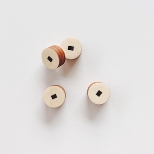 Könnte beinhalten: Vier kleine, runde Holzobjekte mit hellbrauner Farbe und einem dunkelbraunen Rand. Jedes Objekt hat einen quadratischen, schwarzen Einsatz in der Mitte. Die Objekte sind auf einer weißen Oberfläche verstreut.