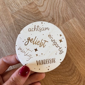 Könnte beinhalten: Eine runde Holzplakette mit den Worten "achtsam", "geliebt", "einzigartig" und "wunderbar" in einer kursivschriftlichen Schrift. Die Plakette ist mit kleinen Sternen verziert.