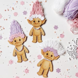 Peut inclure: Trois poupées trolls en bois avec des cheveux en fil rose, violet et blanc. Chaque poupée porte une couronne de fleurs et un collier de perles avec un nom. Des fleurs décoratives et des écheveaux de fil sont dispersés.