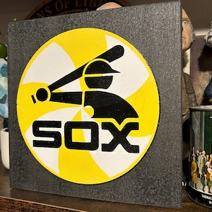 May include: A square wooden sign featuring the Chicago White Sox logo. The logo is a black silhouette of a batter swinging a bat, set against a yellow and white circular background. The word "SOX" is in white block letters.