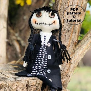 May include: A handmade rag doll with black hair, a black coat, and a white and black checkered dress. The doll has a stitched smile and large brown eyes. The doll is sitting on a tree branch. The text "PDF pattern & tutorial" is in a circle on the image.