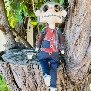 May include: A handmade fabric doll with gray yarn hair, button eyes, and stitched mouth features. The doll wears a red and blue plaid long-sleeve shirt with three buttons, a small dark blue pocket, and dark blue denim pants with a heart button.