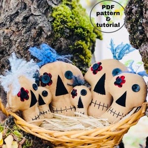 Op de afbeelding: Vier handgemaakte stoffen schedelpoppen in een rieten mand. De poppen zijn beige met zwarte ogen en monden, en hebben rode en blauwe stoffen accenten. De poppen zijn gerangschikt in een nest van stro.