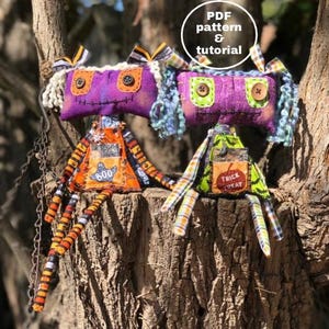 Puede incluir: Dos muñecas de trapo de Halloween hechas a mano con cuerpos de tela morada, piernas rayadas de naranja y negro y ropa de tela colorida. Una muñeca tiene un parche de "BOO" en su vestido y la otra tiene un parche de "TRICK TREAT". Las muñecas están sentadas en un tronco de árbol.