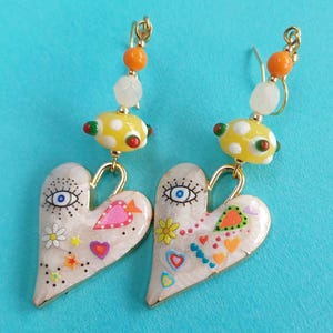 May include: A pair of quirky heart-shaped earrings. The earrings have a cream base with colourful painted details, including eyes, flowers, and hearts. They have gold-coloured hooks and are adorned with yellow and orange beads.