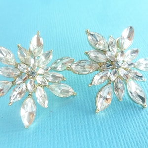 May include: A pair of gold tone earrings with clear crystal flower designs. Each earring features a cluster of marquise-shaped crystals arranged in a starburst pattern.