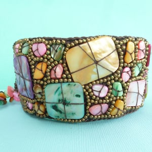 Könnte beinhalten: Ein braunes gewebtes Armband mit einem bunten Mosaikdesign. Das Armband besteht aus kleinen, unregelmäßig geformten Muschel- und Perlenstücken. Die Farben umfassen Pink, Grün, Gelb und Lila.