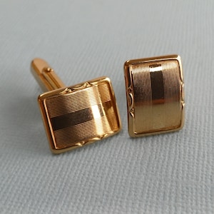 Könnte beinhalten: Ein Paar goldfarbene Manschettenknöpfe. Jeder Manschettenknopf ist rechteckig mit einem strukturierten, gestreiften Design und einem dekorativen Rand. Die Manschettenknöpfe haben ein klassisches, formelles Aussehen.