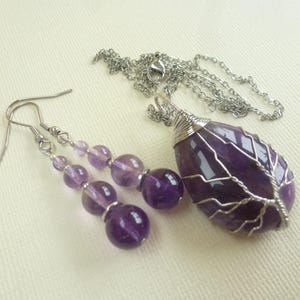 Op de afbeelding: Een sieradenset met een hangerketting en bijpassende oorbellen. De hanger is een druppelvormige paarse steen gewikkeld in zilverdraad, hangend aan een zilveren ketting. De oorbellen hebben paarse kralen. De set ligt op een crèmekleurig oppervlak.