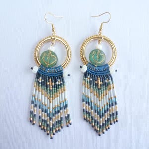 May include: A pair of handmade earrings featuring gold-colored hoops, teal and gold beaded accents, and long fringe. The earrings have a unique design with a combination of blue, white, and gold beads, creating a bohemian style.