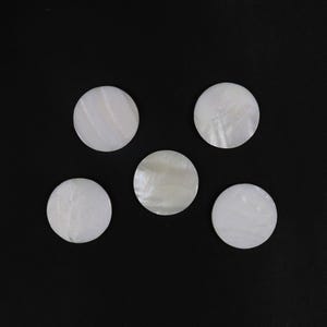 May include: Five round, white mother-of-pearl discs are arranged against a black background. The discs have a pearlescent sheen, reflecting light. The discs are likely for jewelry or craft projects.