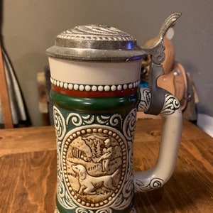 May include: A ceramic beer stein with a green, red, and white design. The stein features a hunting scene with a dog and a hunter. The text "Point English Setter" is visible on the stein.