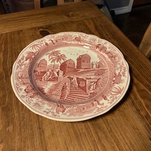 May include: A pink and white ceramic dinner plate with a detailed scene of a village with buildings and people. The plate has a scalloped edge and a white rim.