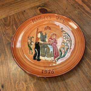 May include: A brown ceramic plate featuring a painted scene of a mother and child. The plate is inscribed with the words "Mother's Day" and the year "1976".