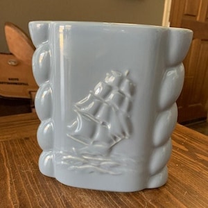 May include: A light blue ceramic vase with a raised relief of a sailboat. The vase has a rectangular shape with rounded edges and a textured, bubble-like design on the sides. The sailboat is depicted with sails billowing in the wind.