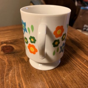 May include: White ceramic mug with a floral design featuring orange, yellow, green, and blue flowers. The mug has a handle and a pedestal base.