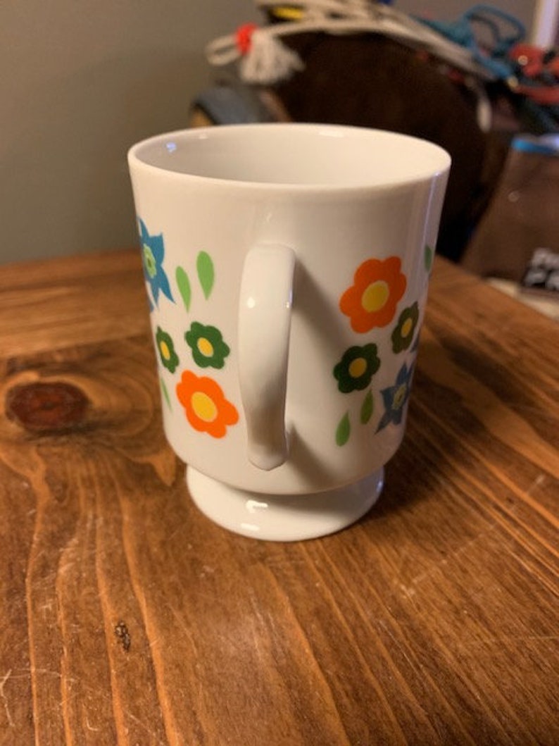 May include: White ceramic mug with a floral design featuring orange, yellow, green, and blue flowers. The mug has a handle and a pedestal base.