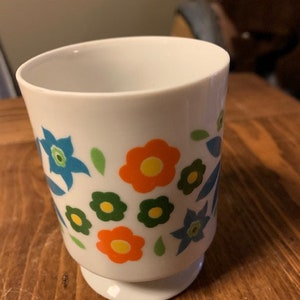 May include: White ceramic mug with a floral design featuring blue, orange, green, and yellow flowers. The mug has a wide base.