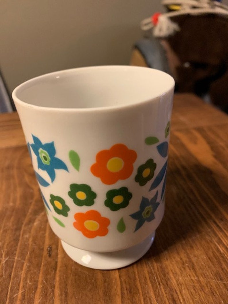 May include: White ceramic mug with a floral design featuring blue, orange, green, and yellow flowers. The mug has a wide base.
