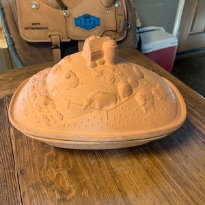 Puede incluir: Una cazuela de terracota con un diseño en relieve de una escena de granja, que incluye un granero, animales y plantas. La cazuela es de forma ovalada y tiene una tapa.
