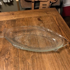 Könnte beinhalten: Eine klare, fischförmige Servierplatte aus Glas mit strukturierter Oberfläche. Die Platte steht auf einem Holztisch.