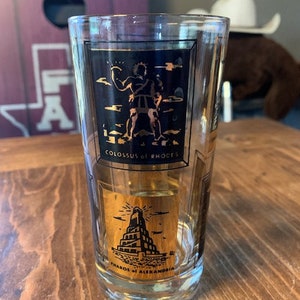 Può includere: Un bicchiere di vetro trasparente con bordi dorati e immagini in bianco e nero del Colosso di Rodi e del Faro di Alessandria. Il bicchiere ha il testo "Colossus of Rhodes" e "Pharos of Alexandria" stampato su di esso.