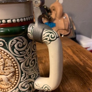 May include: A ceramic beer stein with a green and white floral design. The stein has a silver lid and a white handle.