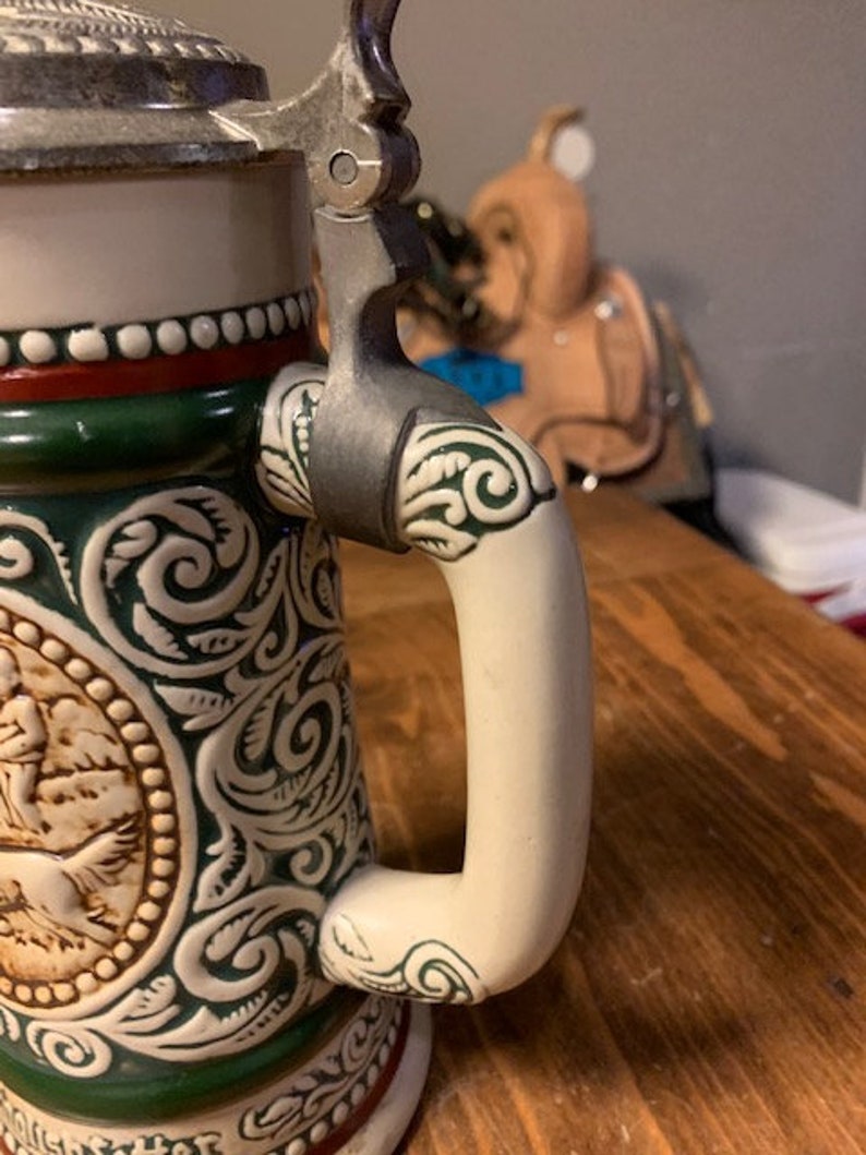 May include: A ceramic beer stein with a green and white floral design. The stein has a silver lid and a white handle.