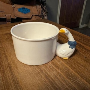 May include: White ceramic mug with a goose-shaped handle. The goose is white with a blue scarf and yellow beak.