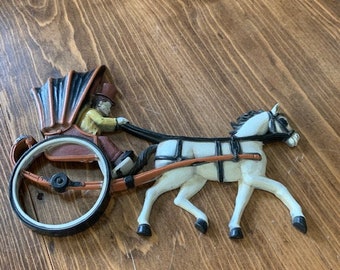 May include: A white horse pulling a brown carriage with a person in a yellow jacket and black top hat. The carriage has a black and brown canopy. The horse and carriage are made of painted metal.
