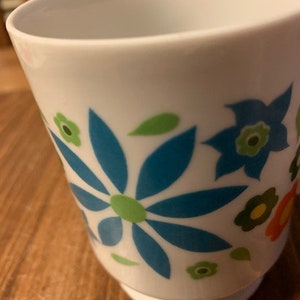 May include: White ceramic mug with a blue, green, and orange floral design. The flowers are stylized and have a retro look.