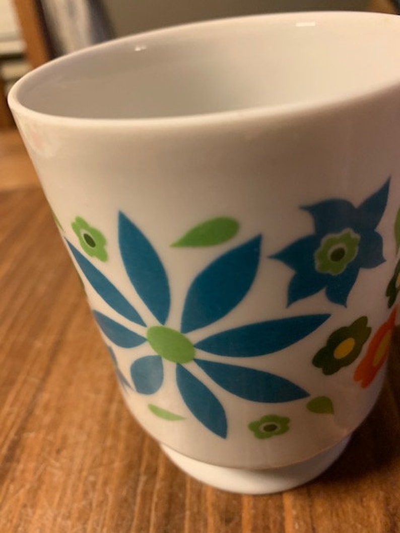 May include: White ceramic mug with a blue, green, and orange floral design. The flowers are stylized and have a retro look.