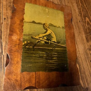 Może przedstawiać: Drewniana tabliczka vintage z obrazem w ramie przedstawiającym osobę wiosłującą łódką po jeziorze. Obraz jest w odcieniach zieleni i brązu.
