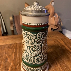 May include: A ceramic beer stein with a green and white floral design. The stein has a pewter lid and a red and white band around the top. The text "At Point" is visible on the bottom of the stein.