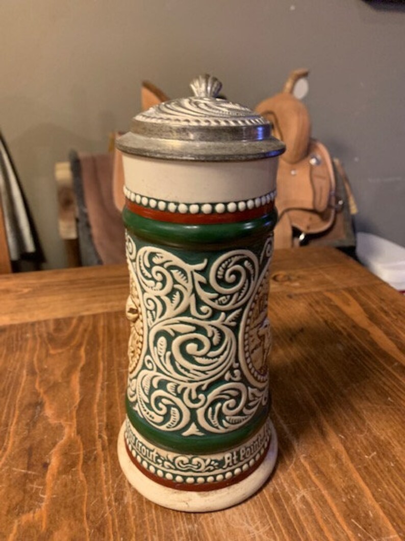 May include: A ceramic beer stein with a green and white floral design. The stein has a pewter lid and a red and white band around the top. The text "At Point" is visible on the bottom of the stein.