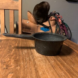 May include: A black speckled enamel saucepan with a black handle. The saucepan is sitting on a wooden table.