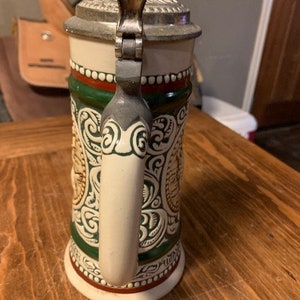 May include: A white ceramic beer stein with a green and red band around the middle. The stein has a detailed floral design and a metal lid with a handle.