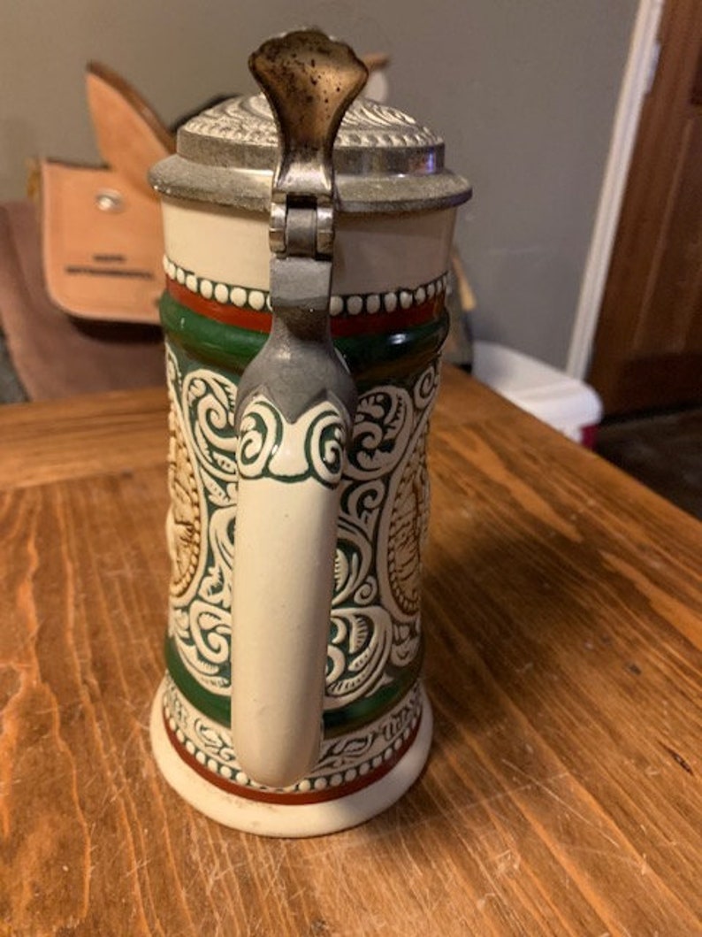 May include: A white ceramic beer stein with a green and red band around the middle. The stein has a detailed floral design and a metal lid with a handle.