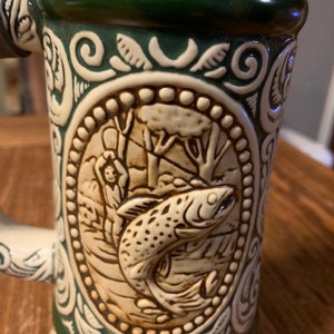 May include: A ceramic beer stein with a green and white design. The stein features a detailed image of a fish and a person fishing. The stein has a red, white, and green band around the top.