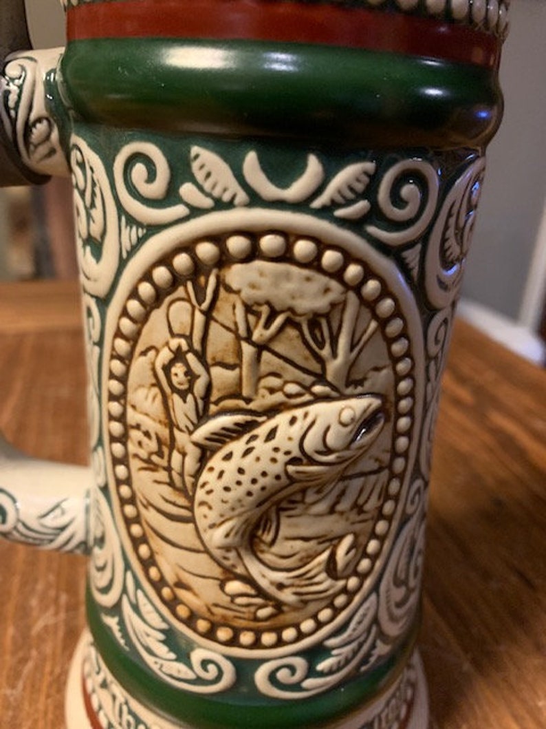 May include: A ceramic beer stein with a green and white design. The stein features a detailed image of a fish and a person fishing. The stein has a red, white, and green band around the top.