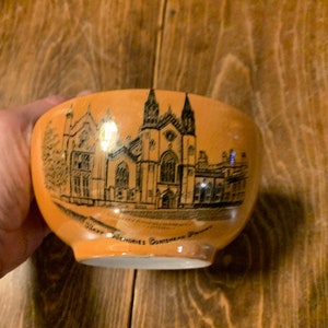 May include: A small, orange ceramic bowl with a black and white illustration of a building. The bowl has the words "Wills Antiques, Constantia, Pa." printed on the bottom.