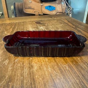 Puede incluir: Un plato de vidrio rojo oscuro con un diseño acanalado y bordes redondeados. El plato tiene dos pequeñas asas a cada lado.