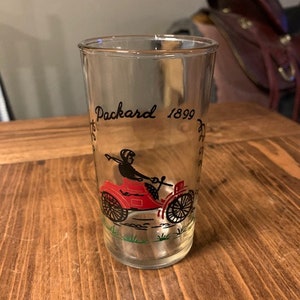 May include: A clear glass with a black and white illustration of a red car with a person driving. The text "Packard 1899" is printed on the glass.