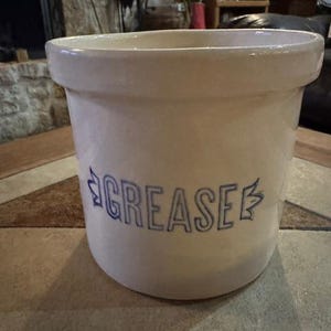 May include: Off-white ceramic grease jar with a wide opening and a slightly flared rim. The word "GREASE" is printed in blue, stylized lettering on the front. The jar is cylindrical and sits on a tiled surface.