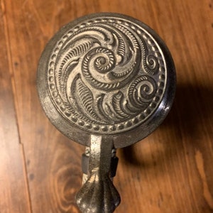 May include: A silver beer stein with a decorative lid featuring a swirling, embossed design. The lid has a small, ornate handle.